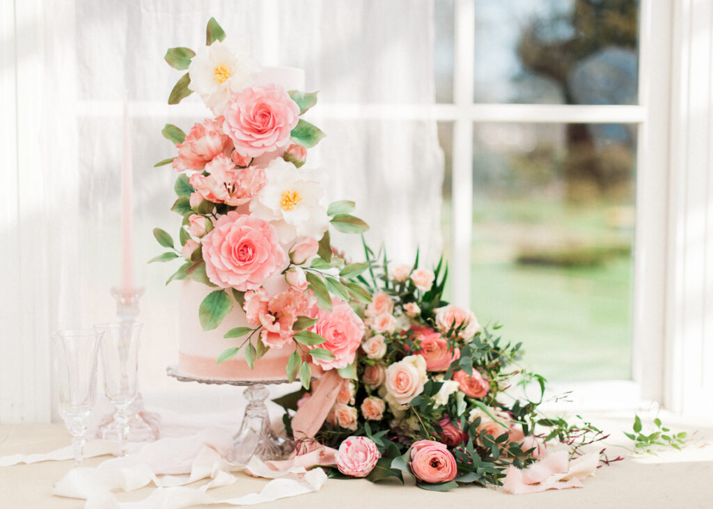 a pink 3 tiered wedding cake with white and pink camellia sugar flowers, pink tulips and green foliage
